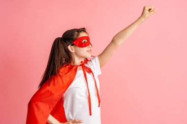 Süper kahraman kostümlü, kırmızı pelerinli ve maskeli güzel bir kız ne kadar güçlü olduğunu gösteriyor. Pembe arka planda soyutlanmış. Süper kahraman oyunu oynayan şirin bir bebek. Kız Gücü konsepti.