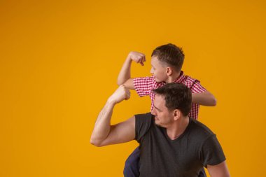 Dost canlısı bir adam ve elleri havada küçük bir çocuk kaslarını ve güçlerini gösteriyor. Baba ve oğul stüdyoda sarı arka planda poz veriyorlar..