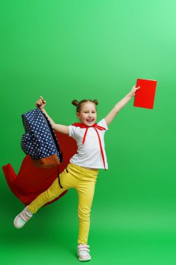 Kırmızı süper kahraman pelerinli mutlu kız öğrenci elinde okul kitabı ve sırt çantasıyla kameraya gülümsüyor ve stüdyonun yeşil arka planında poz veriyor. Çocuk öğrenmeye hazır. Okula geri dön.