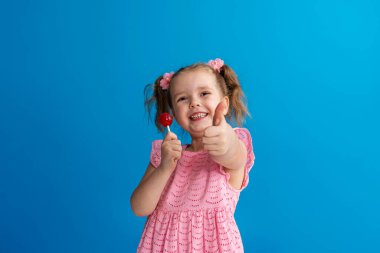 Komik çocuk jest onayını gösteriyor. Çubuğun üzerinde lolipopu olan 4 yaşındaki bir kız komik bir şekilde gülümsüyor ve yeşil arka planda, stüdyoda zevkle poz veren yaramaz küçük bir kız olduğunu gösteriyor. Boşluğu kopyala.