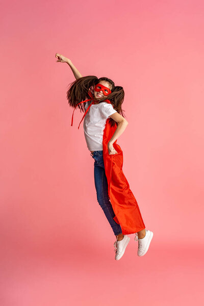 beautiful smiling girl in superhero costume, in red cape and mask shows how strong she is, isolated on a pink background in full growth. Cute baby playing superhero game. concept of Girl Power.