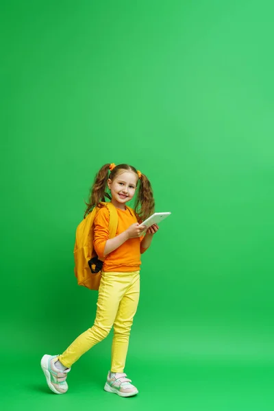 电子学习 可爱的女学生 手里拿着两个马尾辫 背着背包和平板电脑 站在绿色的背景上 孩子笑着看着镜框 推荐远程在线学校 — 图库照片