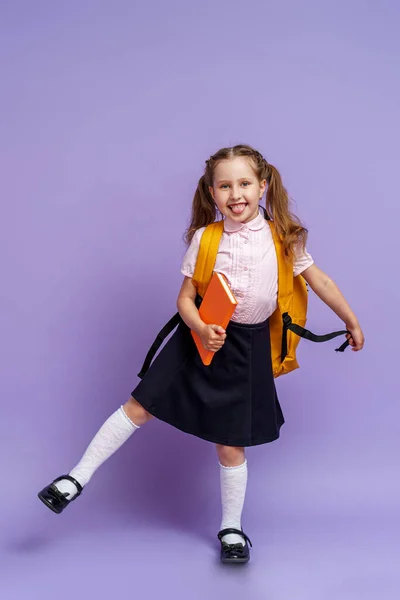 可爱的快乐的孩子穿制服 把他的腿抬起来 在紫色的背景上跑 背着背包的孩子 小女孩已经准备好上学了 可以追溯到概念学校的动态图像 假期开始 — 图库照片