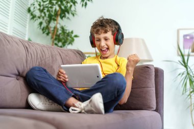 Kulaklığı ve tableti olan mutlu çocuk evde koltukta oturuyor. Çocuk evde bilgisayar tabletinde müzik dinleyerek öğrenmekten mutlu. Yeni çocuk öğrenme uygulaması. Aletlere bağımlılık