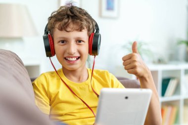 Kulaklıklı ve tabletli mutlu çocuk evde, kanepede oturuyor. Çocuk evde bilgisayar tabletinde müzik dinleyerek öğrenmekten mutlu. Çocuklara öğretmek için yeni bir uygulama.