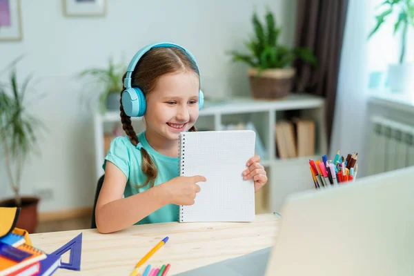 ヘッドフォンをしている少女はノートパソコンを使って先生とビデオ通話をします 子供は遠隔で学ぶことができます 女子高生は喜んで知識をもらう Education距離ホームラーニング — ストック写真