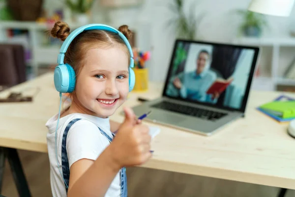 ヘッドフォンをしている少女はノートパソコンを使って先生とビデオ通話をします 子供は遠隔で学ぶことができます 女子高生は喜んで知識をもらう Education距離ホームラーニング — ストック写真