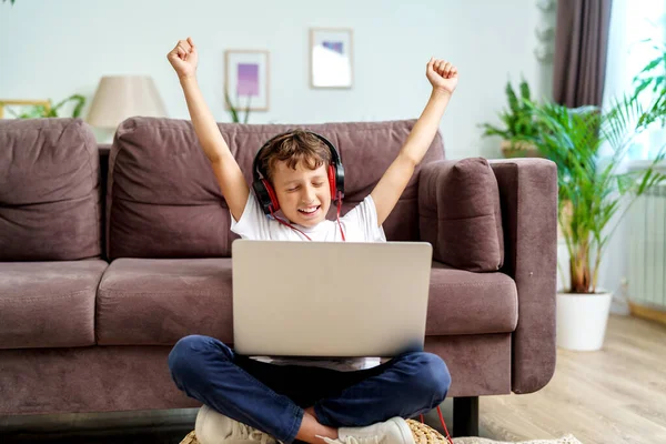 ヘッドフォンを持つ幸せな巻き毛の少年は彼の手を上げ 笑いと喜び ソファの上に足を組んで座って ラップトップでビデオゲームを受賞することを祝います — ストック写真
