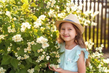 Sevimli küçük kız, güneşli yaz bahçesinde yasemin çiçeklerini kokluyor. Kafasında şapkası ve açık çizgili elbisesi olan çocuk güzel çiçeklerden hoşlanır. Romantik konsept şefkat, çocukluk.