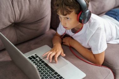 Kulaklıklı, dizüstü bilgisayarı koltukta uzanan çocuğun yakın çekimi, çocuk evde ders dinleyerek, bilgisayarında çalışmaktan mutlu. Çocuklara öğretmek için yeni başvuru formu. Beyaz ekran