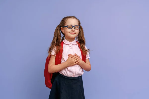 戴眼镜 身穿校服 背着背包 双手放在胸前 与紫色背景工作室隔离的快乐小女孩 微笑迷人的心满意足的小可爱表现出对学习的热爱 — 图库照片