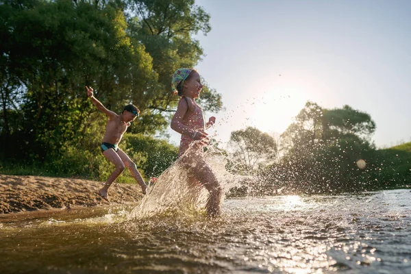 Happy children run from the shore into the water. Summer children\'s vacation on shore of a lake or river. boy and girl jump into water, swim and splash around at sunset. Active holidays. Dynamic image