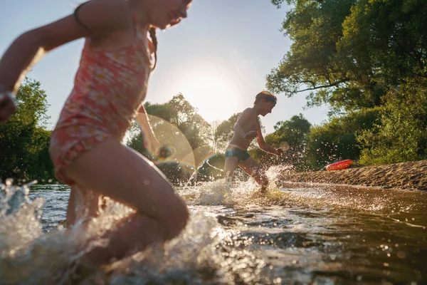 Happy children run from the shore into the water. Summer children's vacation on shore of a lake or river. boy and girl jump into water, swim and splash around at sunset. Active holidays. Dynamic image