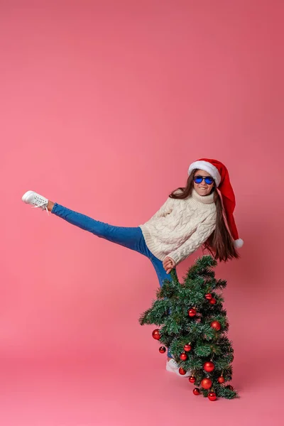 身穿针织毛衣 戴着太阳镜 头戴圣诞帽 头戴红球装饰的圣诞树的快乐少女 在粉色的背景下行走 为圣诞节做准备 — 图库照片