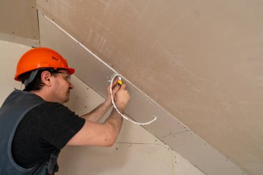 Alçıpan dikişlerini işleme ve budama işlemi. Tulumlu ve turuncu kasklı bir erkek inşaat işçisi alçıpanın kenarlarını keser ve hizalar. Onları inşaat alanında macuna hazırlar..