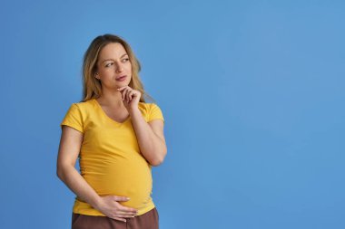 Çekici, düşünceli hamile bir kadın, midesini okşuyor ve mavi arka planda dikilirken düşünceli bakışlarını kaçırıyor. Çocuk bekleyen bir kadın bir seçim yapar. Boşluğu kopyala