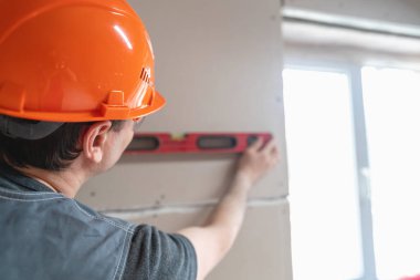 Alçıpan dikişlerini işleme ve budama işlemi. Tulumlu ve turuncu kasklı bir erkek inşaat işçisi alçıpanın kenarlarını keser ve hizalar. Onları inşaat alanında macuna hazırlar..