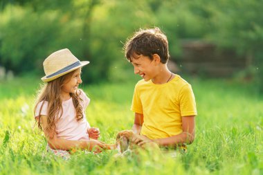 Mutlu çocuklar, bahçedeki güneşli bir günde çimlerde oturan bir tavşanı havuçla okşayıp besliyorlar. Dikkat et ve evcil hayvana bak. Tüylü tavşan onların arkadaşı..