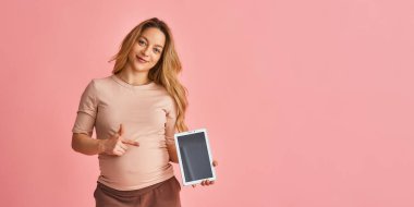 Çekici hamile kadın elinde tablet bir bilgisayar tutuyor ve parmağını alet ekranına doğrultuyor. Müstakbel anne pembe arka planda duran yeni bir başvuru ilanı veriyor. Boşluğu kopyala.