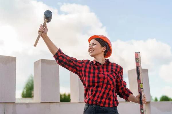 young female construction worker in protective helmet, holding mallet and construction level in her hands, stands against sky and raises her hands with construction tools. Copy space