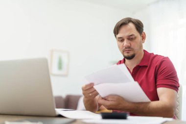 Genç erkek işadamı evde dizüstü bilgisayarında belgelerle çalışıyor. Ev ofisinde çalışıyor. İnternette okuyor. İnternet bağımlılığı mı yoksa bütün gün çalışan biri mi? Düşünceli bir adam ev ofisinde çalışıyor.