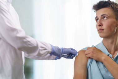 Doktor, maske takan yetişkin bir delikanlıya omzuna iğne yapıyor. Yetişkinlerin koronavirüse karşı aşılanması. Covid-19 'a karşı aşı. Virüse karşı koruma. Kapatın. Boşluğu kopyala