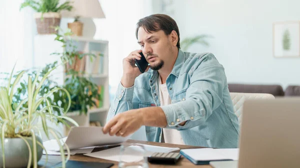 young male businessman works at home with documents on laptop, works from home office, studies online. positive adult, solves questions on phone, works all day. thoughtful person works in home office