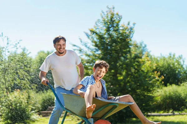 happy, joyful boy, pre-adolescent age, is having fun and laughing while riding garden wheelbarrow, which is pushed by his father in backyard garden on warm sunny day. Active family games in fresh air