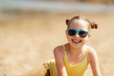 Güneş gözlüklü mutlu küçük bir kızın portresi. Yazın güneşli bir günde kumsalda yatıyor. Bir çocuk göl kıyısında yaz tatilinin tadını çıkarıyor. Aktif yaz tatili