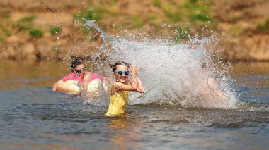 Mutlu çocuklar suda koşup oynuyorlar. Gölün veya nehrin kıyısında çocukların yaz tatili. Bir erkek ve bir kız gün batımında suya atlıyor, yüzüyor ve su sıçrıyor. Aktif eğlence. Dinamik resim