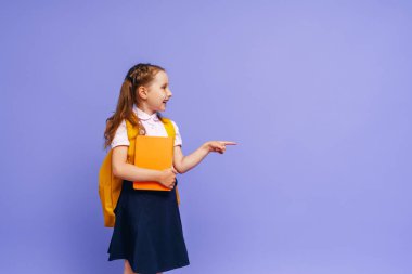 Güzel, mutlu bir bebek. Parmağıyla mor arka planı işaret ediyor. Sırt çantalı çocuk. Küçük kız okula gitmeye hazır, kavramsal okula geri dönüyor. Tatil başlıyor..
