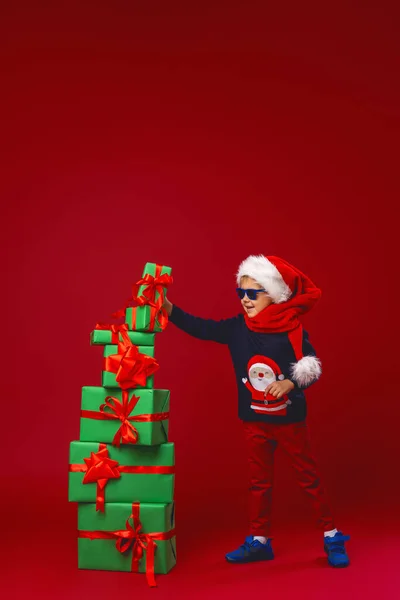 在工作室里 戴着太阳镜 头戴圣诞礼帽的小男孩在红色背景上建造了一个圣诞礼品盒金字塔 孩子高兴地微笑着 并收到了许多圣诞礼物 — 图库照片