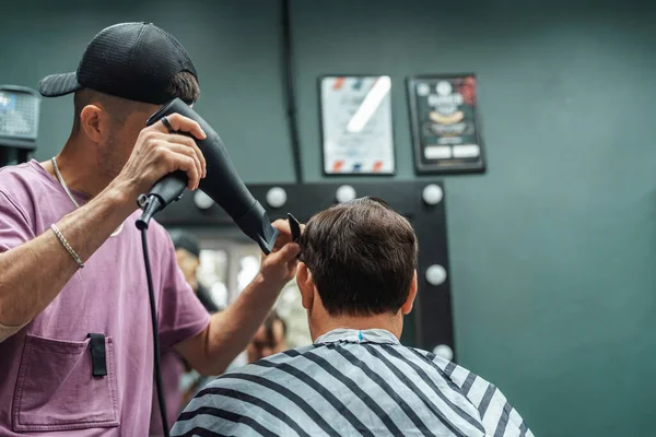 young barber guy dries hair bearded man sitting in chair in barber shop with hair dryer. man cut his hair with scissors and comb. Men\'s haircuts and beard shaving. Haircut and edging of eyebrows.