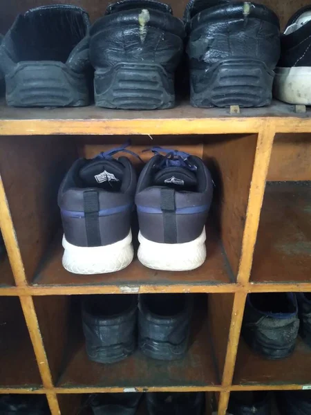 Bir çift lacivert ve beyaz spor ayakkabısı, yukarıdaki ve aşağıdaki raflarda çeşitli koyu renkli ayakkabılarla çevrili ahşap bir ayakkabı rafının orta rafında duruyor.