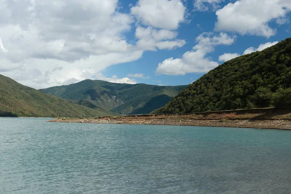 Dağlık bir göl manzarası, kayalık bir kıyı şeridi ve bulutlu mavi gökyüzünün altındaki ormanlık tepeler..