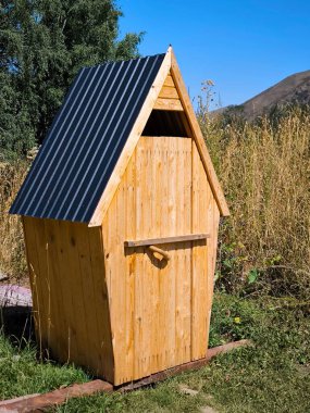 Klasik, açık hava tuvaleti, açık ahşaptan yapılmış, çatısı siyah kıvrımlı metalle kaplı. Tuvalet kırsal bir alanda, bir yazlık evde ya da bir kamp alanında bulunur.. 