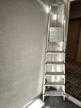 Beş basamaklı alüminyum ya da metal merdiven kalın, desenli alçıdan bir duvara dayanır..