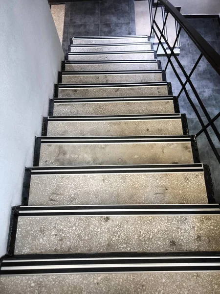 Düz bir merdiven boşluğunun dibinden bir manzara. Basamaklar açık terrazzo ya da betondan ve belirgin siyah beyaz kayma önleyici şeritlerden yapılmış. Siyah metal parmaklıklar sağda görünüyor..