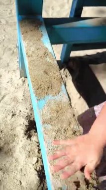 Çocuk parkında birlikte oynayan iki çocuğun elleri. Islak kumu serpmek ve tırmıklamak için mavi ahşap bir kiriş kullanıyorlar. Paylaşılan oyun, tasasız çocukluk, arkadaşlık.