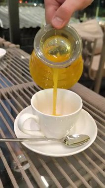 Bir el, parlak turuncu deniz dikenli çaydanlıktan beyaz bir porselen fincana zengin bir portakal çayı döker. Fincan, gümüş bir kaşıkla örülmüş bir masanın üzerinde duruyor..