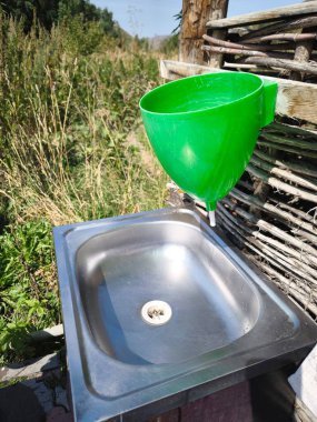 Paslanmaz çelik bir leğen ve hasır bir çite bağlı parlak yeşil plastik bir el havuzu olan basit bir açık hava yıkama tezgahı. Tipik bir kırsal ya da taşra tarzı el yıkama istasyonu.