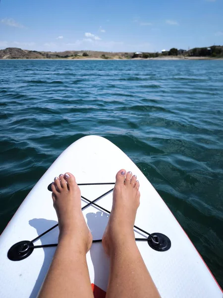 Hafif çalkantılı mavi sularda yüzen beyaz bir SUP tahtasının üzerinde oturan bir insanın bacakları, sakin bir yaz tatili ve sudaki macera atmosferini aktarır..