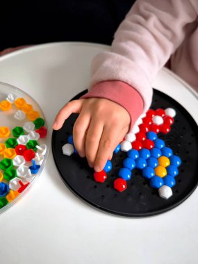 Yuvarlak bir kara tahta üzerinde altıgen mozaik oluşturmakla meşgul bir çocuk eli. Çok sayıda renkli parçanın bulunduğu bir konteynır yakınlarda duruyor..