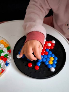 Yuvarlak bir kara tahta üzerinde altıgen mozaik oluşturmakla meşgul bir çocuk eli. Çok sayıda renkli parçanın bulunduğu bir konteynır yakınlarda duruyor..