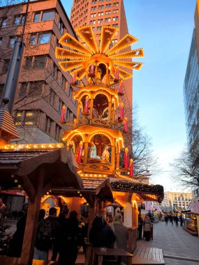 Berlin, Almanya - 2 Aralık 2025: Noel pazarında sıcak sarı ışıklar ve kırmızı mumlarla aydınlatılmış çok katmanlı uzun bir Noel piramidi yapısı.