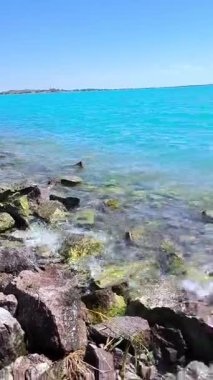 Büyük kayalıkları ve temiz dalgaları olan bir sahil bölgesinin fotoğrafı. İskele vinci olan bir sanayi manzarası ufukta görünüyor..