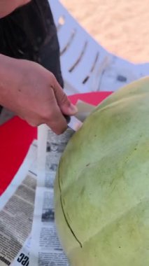 Çok büyük, açık yeşil bir karpuzu dilimliyorum. Bir insan eli, derme çatma bir gazete tabanına dayanan meyvenin çevresini kesmek için büyük bir bıçak kullanır..