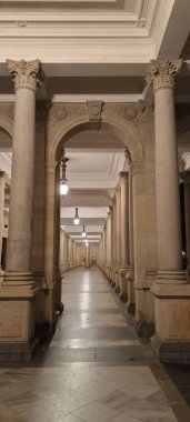 Karlovy Vary, Çek Cumhuriyeti-30 Aralık 2024: Geceleri boş Mlyn Colonnade 'nin görkemli manzarası. Korint sütunları, klasik mimari, sıcak akşam aydınlatmaları ve perspektif bir sükunet ve lüks atmosfer yaratıyor.