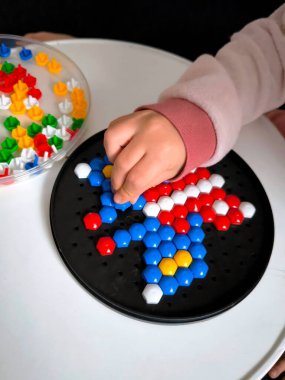 Yuvarlak bir kara tahta üzerinde altıgen mozaik oluşturmakla meşgul bir çocuk eli. Çok sayıda renkli parçanın bulunduğu bir konteynır yakınlarda duruyor..