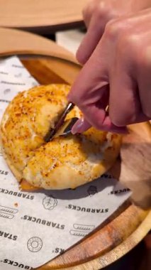 Bitterfeld, Almanya - 23 Aralık 2025: Bir kadın elleri, bir kahve dükkanının içindeki ahşap bir tahtanın üzerinde gevrek bir kabuk ve dolgu ile lezzetli bir yuvarlak sandviç kesti..